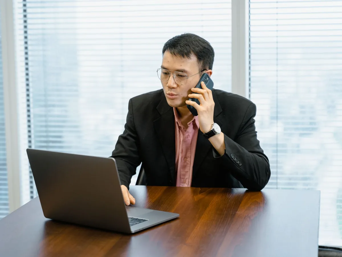 Nam giám đốc người châu Á mặc vest đen, sơ mi hồng, vừa nghe điện thoại vừa làm việc trên laptop trong phòng họp sáng.