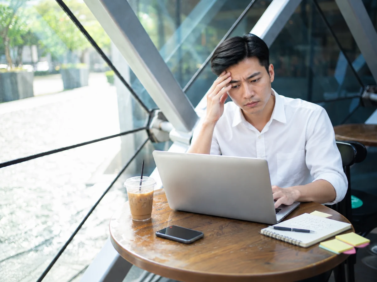 Nam freelancer trẻ người Việt mặc sơ mi trắng, tay đỡ trán, vẫn gõ laptop bên bàn gỗ tròn đầy giấy nhớ, sổ việc và ly cà phê dở trong quán cà phê vách kính.
